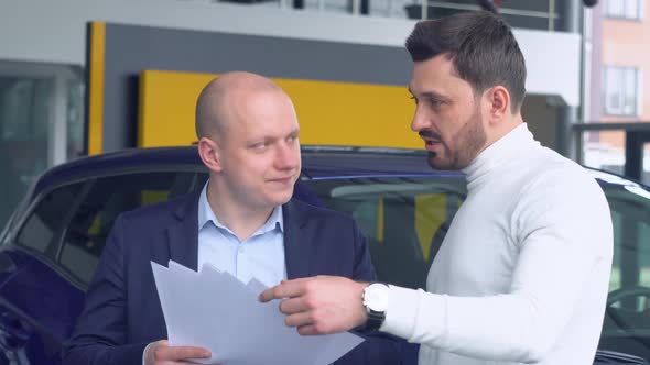 Professional Salesman Smiling Showing a New Car at the Dealership to His Male Customer