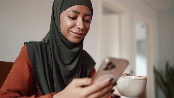 Young Muslim woman drinking tea and typing by phone