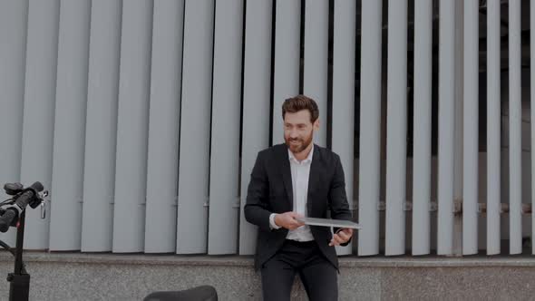 Modern Businessman in a Suit Finishes Work with a Laptop on the Street and Leaves on a Scooter