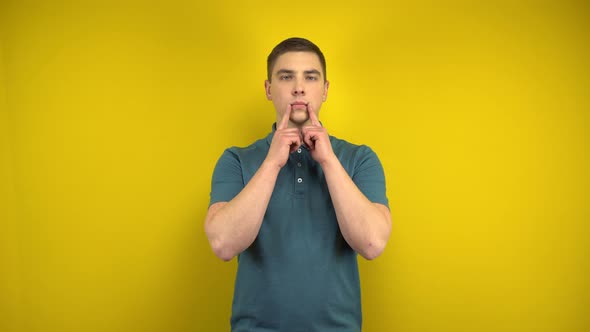 A Young Man Makes a Sad Smile on Her Face with His Fingers on a Yellow Background. Man in a Green