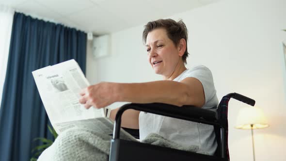 Senior Woman Sitting in a Wheelchair and Reading a Newspaper at Home