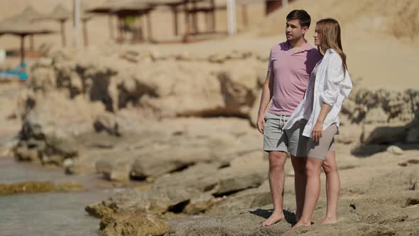 a Man in a Pink Tshirt and a Woman with Long Hair in a White Shirt are Standing Looking at the Sea