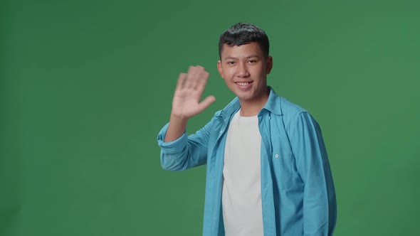 The happy young Asian man waving hand while standing on green screen in the studio