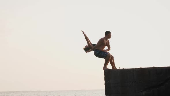 Young Woman and Man Synchronously Doing Trick Jump From a Pier Into the Sea During Beautiful Sunrise