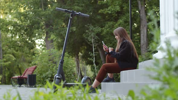 Woman is Sitting on Steps Park Next to an Electric Scooter and Typing Something in Her Phone Side