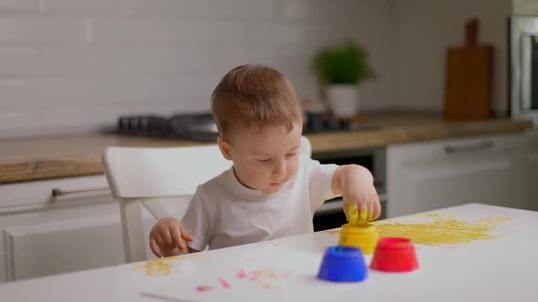 Cute Little Baby Boy Sitting Behind Table and Drawing with Fingers Covered in Colorufl Paint