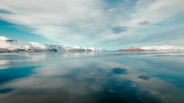 A wide shot hyper lapse, flying over calm water towards a snowy mountain scape with soft clouds roll