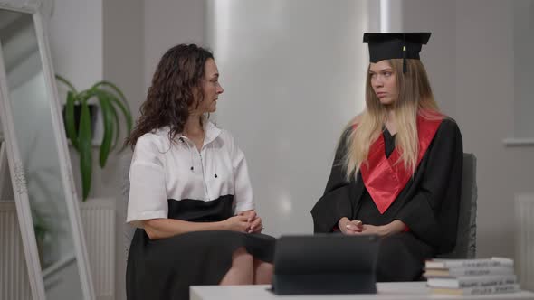 Serious Caucasian Mature Mother Talking with Young Graduate Daughter Sitting Indoors at Home