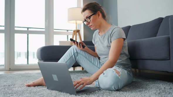 Casually Dressed Woman Sitting on Carpet with Laptop and Smartphone and Working in Cozy Room