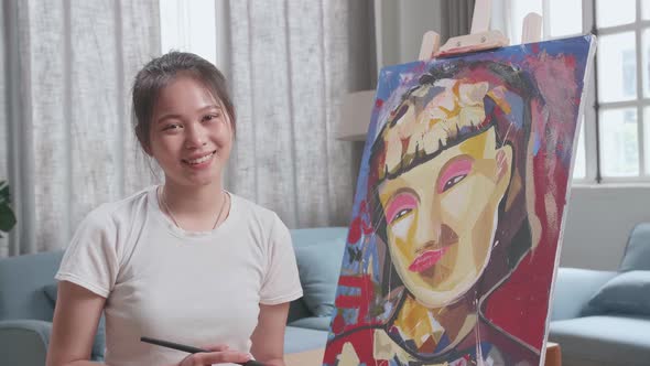 Asian Girl Sitting And Holding Paintbrush And Smiling After Finish Painting A Girl On The Canvas