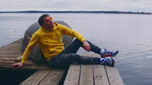 Young Man in Bright Glasses and Hoodie Sits Leaning on Boat on Wooden Pier and Looking Away on