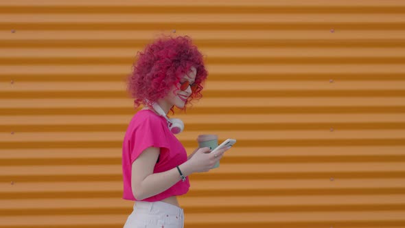 A Pink Curly Teenager is Walking and Using a Smartphone Outdoors alt