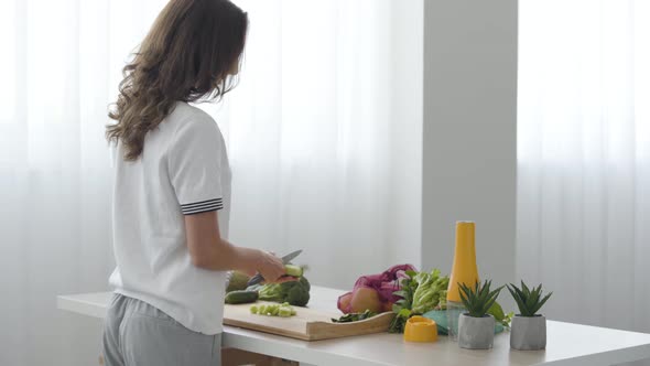 Skill Girl Cuts a Cucumber with Knife While Standing at a Table in the Kitchen at Home