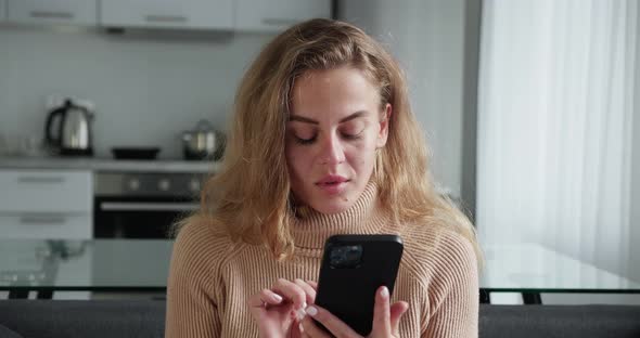Curly Blonde Woman Looking in Smartphone Typing Message or Surfing News Sitting in the Cozy Sofa