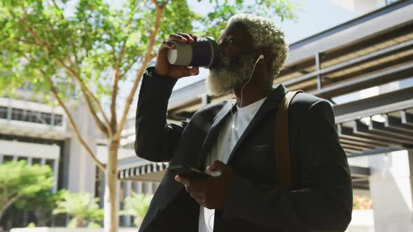 African american senior man drinking coffee and using smartphone in corporate park