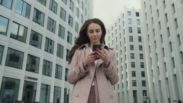 Cheerful young Caucasian woman standing in city street and texting on mobile phone with smile.