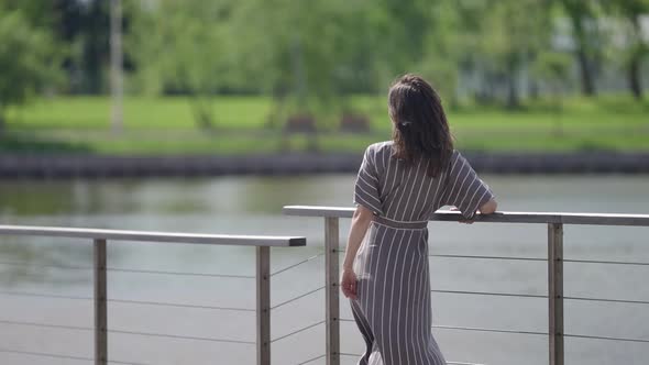 a Brunette in a Striped Dress Stands at the Railing of a Bridge Over the River on a Windy Sunny Day