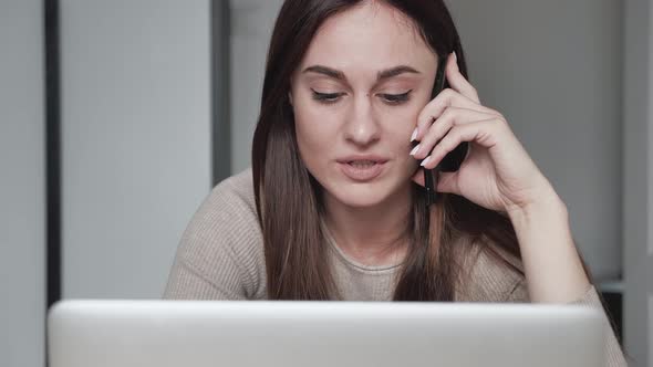 An Attractive Young Woman Freelancer Speaks on a Smartphone at a Computer and Looks at the Monitor