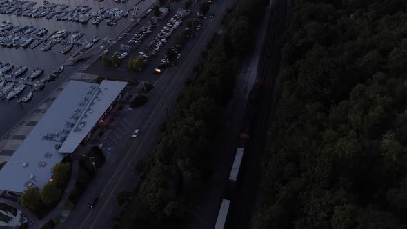 Aerial Of Train Passing Cars By Ocean Boat Marina At Dusk alt