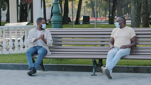 Two African American Men in Facemask Sitting on Bench Keeping Distance Discussing Problems of