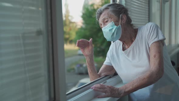 Lonely Senior Woman in Mask Stands By Window and Waves Her Hand to Family Who Came Visit Her at