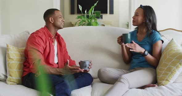 Happy biracial couple sitting on sofa with coffee and talking