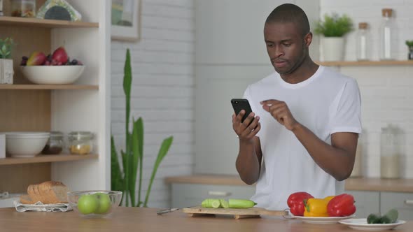 Young African Man Using Smartphone While Cutting Vegetables in Kitchen