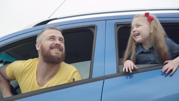 Side View of Blue Car with Smiling Happy Young Father and Little Daughter Sticking Out Windows