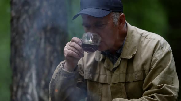 Elderly Forester is Resting in Forest and Drinking Tea Near Campfire Halt in Woodland