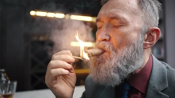 Zoom Out Portrait of Bearded Mature Businessman Smoking Cuban Cigar with Burning Match Sitting at