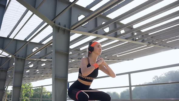 Young Woman in Earphones which Doing Squat Exercises Using Fabric Booty Band Outdoors