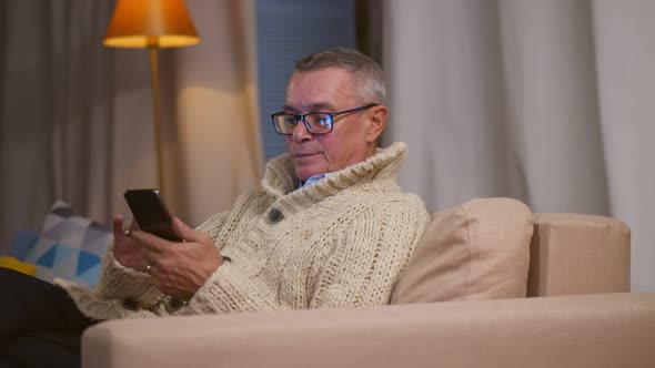 Senior Man Using Smartphone Browsing Internet Sitting on Couch at Home