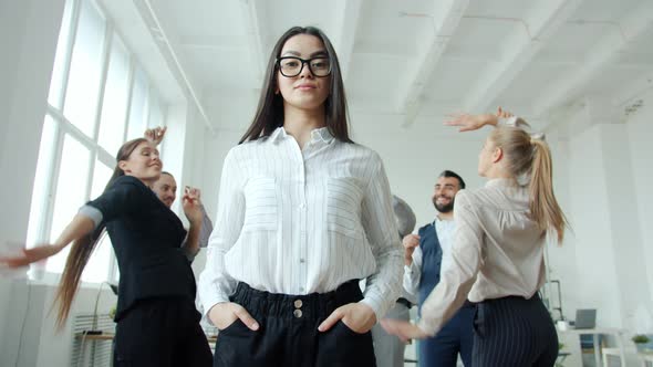 Young Asian Businesswoman with Serious Face Standing in Office Where People Men and Women Dancing