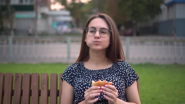 A Young Pregnant Girl is Eating a Burger While Sitting on a Bench in the Park