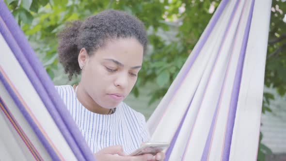 Close-up Face of Young African American Woman Sitting in the Hammock, Relaxing in the Garden