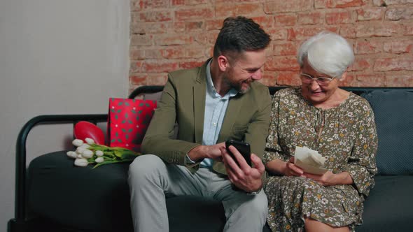 Greyhaired Elderly Pensioner Woman Sitting on a Sofa and Looking at Her Adult Millennial Son's