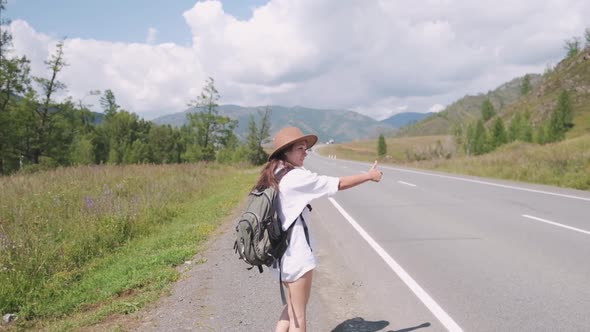 Attractive Brunette Girl in a Hat with a Backpack Travels in the Mountains Hitchhiking, Votes the