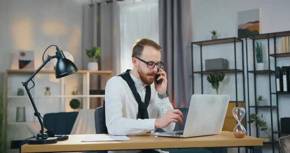Office Worker in Glasses Talking on Phone while Working on Computer in Good-Decorated Home Office