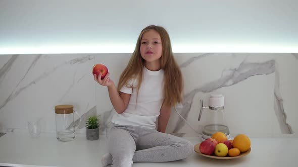 Little Girl with Long Fair Hair Eating Red Apple in Bright Modern Kitchen While Sitting on a Counter