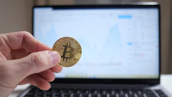 Male Hand Holding Cryptocurrency Coin Over Laptop Background Showing Graph