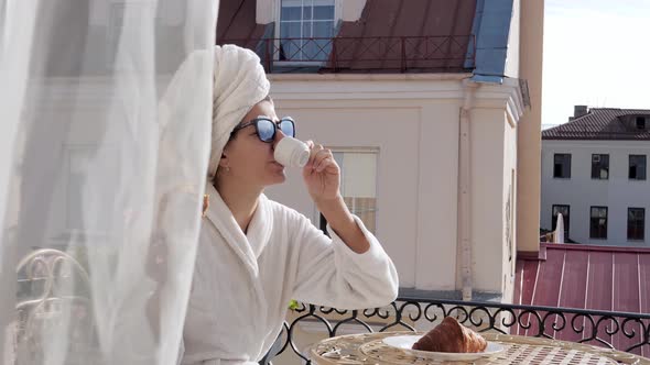 In Morning Woman In Bathrobe On Balcony Has Breakfast With Coffee And Croissant