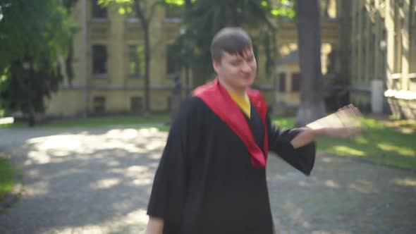 Portrait of Excited Young Man in Graduation Toga Dancing in Sunshine at College Campus Looking at