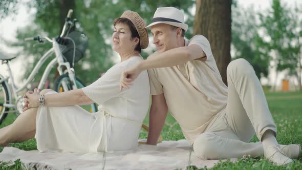 Elegant Mid-adult Caucasian Couple Sitting on Blanket in Summer Park and Chatting. Portrait of Happy