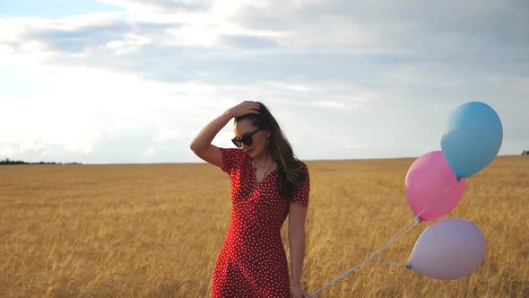 Young Woman in Sunglasses Standing Against the Background of Wheat Field and Holding Balloons in