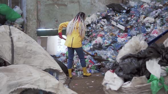 A Rare View of a Girl with Dreadlocks in Yellow Boots Scoops Used Bottles with a Large Duck Shovel