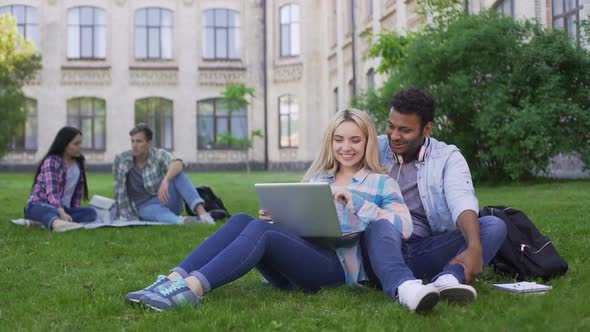 Multi-Ethnic Students Sitting on Grass and Watching Funny Video on Laptop