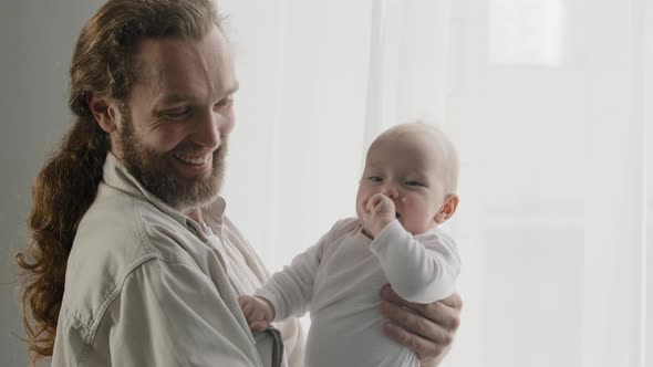 Caucasian Bearded Father Holding Small Happy Baby at Home Standing Near Window Looking at Camera