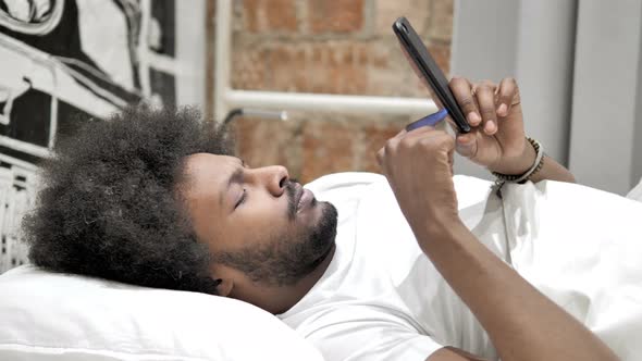 African Man Using Smartphone, Lying in Bed