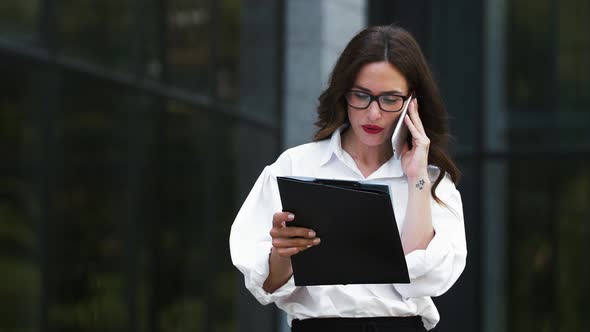 Girl in Glasses and White Shirt Tanding Near Business Center with Papers on Clipboard