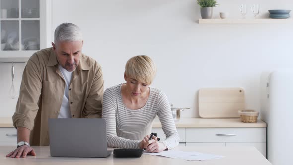 Mature Couple Calculating Expenses Taking Notes Planning Budget In Kitchen